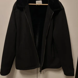 Svart jacka i mockaimitation från Studio Total - Svart jacka från Studio Total med dragkedja och mjuk krage i päls. Kragen samt hela insidan är täkt i fuskpäls, och den yttre sidan består av mockaimitation. Passar bra för kyligare dagar samt till vår och höst.
