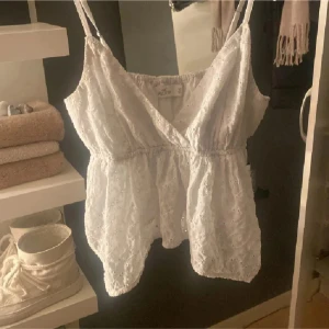 Vit babydoll topp från Hollister - Säljer en söt slutsåld vit topp från Hollister med broderade detaljer och justerbara axelband. Toppen har en omlott-design och elastisk midja för en bekväm passform. Perfekt för varma sommardagar! Använt enstaka gång och köpt på zalando.