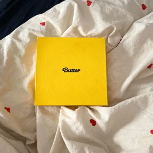 Butter Album Box - Säljer en gul albumlåda med texten 'Butter' på framsidan. Inuti finns klistermärken med färgglada motiv och texter som 'Be Happy' och 'Besties'. Perfekt för fans av musikgruppen.