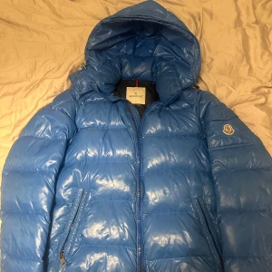 Blå dunjacka från Moncler - Säljer en snygg blå dunjacka från Moncler. Jackan har en glansig finish och är utrustad med en dragkedja framtill. Den har en huva och två praktiska fickor med dragkedja. Perfekt för kyliga dagar! Jackan har är äkta och har bevars fint. Kartologen är borta. Som ni ser på sista bilden har vissa knappar blivit lite blåa. Men absolut ingenting man tänker på! Skick 4/5. Jackan är i stork 5/L men jag har storlek M vanligtvis och använder den!