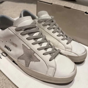 Golden Goose skor - Snygga vita sneakers från Golden Goose med grå stjärndetalj och silverfärgad häl. Skorna har grå snörning och en unik design med en sliten look. Perfekta för en trendig och avslappnad stil. Tillverkade i Italien och KVITTO FINNS! Har haft dem i 3 månader ungefär och dem är varsamt använda 