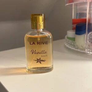 La Rive Vanilla Touch Eau de Parfum - En elegant flaska med gyllene lock innehållande La Rive Vanilla Touch Eau de Parfum. Doften har en söt och varm vaniljton, perfekt för den som älskar mjuka och inbjudande dofter. Flaskan är genomskinlig med svart text och en stilren design.