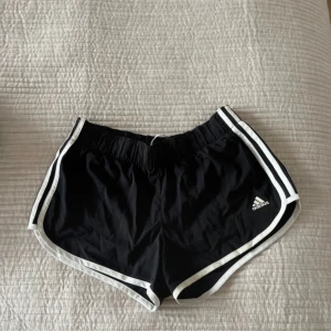 Svarta träningsshorts från Adidas - Säljer ett par svarta träningsshorts från Adidas med vita ränder längs sidorna. Shortsen har en elastisk midja och är perfekta för sport och träning. Adidas-loggan är tryckt på ena benet.