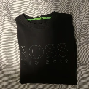 Svart tröja från Hugo Boss - Snygg svart tröja från Hugo Boss med deras logga tryckt på framsidan. Tröjan har en rund halsringning och långa ärmar. 