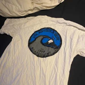 Vit t-shirt från Quiksilver - Säljer en vit t-shirt från Quiksilver med ett stort tryck på ryggen och ett mindre på bröstet. Trycket har en blå och svart design med texten 'Boardriding Est 1969'. Perfekt för en avslappnad stil. Gammal tröja retro/vintage.