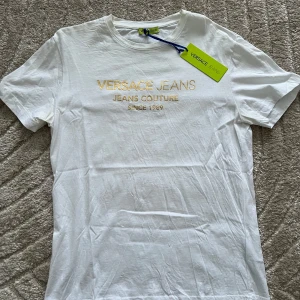 Vit t-shirt från Versace - HELT NYTT! Snygg vit t-shirt från Versace Jeans Couture med guldfärgad text på bröstet. Perfekt för en stilren look. T-shirten har en klassisk rund hals och korta ärmar. Så klart äkta!!