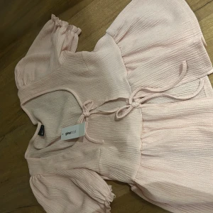 Ljusrosa blus från Gina Tricot - Söt ljusrosa blus från Gina Tricot med korta ärmar och knytning framtill. Blusen har en lätt volangdetalj vid ärmslut och en luftig passform. Den är helt ny med pris lappen kvar.