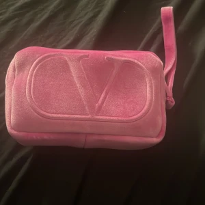 Rosa necessär valentino  - Snygg rosa necessär i sammet med en stor logotyp på framsidan. Den har en praktisk dragkedja och en liten rem för enkel hantering. Perfekt för att förvara smink eller andra småsaker. Använd några få gånger men mest ba legat på sminkbordet 
