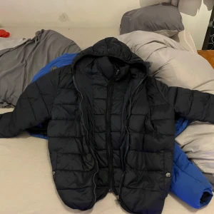 Svart pufferjacka med huva - Säljer en svart pufferjacka med huva och dragkedja. Jackan har långa ärmar och praktiska fickor med knappar. Perfekt för kyligare dagar.