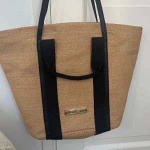 intimissimi beach bag - Snygg beige shoppingväska med svarta handtag och detaljer. Perfekt för en dag på stan eller stranden. Väskan har en stilren design med en metallplatta framtill.