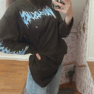 Svart oversized tröja - Womankind - Cool svart tröja med blått tryck på bröstet och ärmarna. Tröjan har en avslappnad, oversized passform och långa ärmar. Perfekt för en casual look.