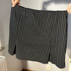 Svart kortkjol med vita ränder - Snygg svart kortkjol med vita ränder. Perfekt för en stilren look. Kjolen har en tight passform och är enkel att matcha med olika toppar. NYSKICK