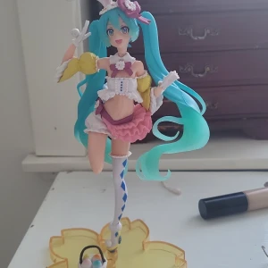 Miku  påsk - En färgglad figur av en karaktär med långt turkost hår och kaninöron. Figuren har en vit och rosa outfit med gula detaljer och står på en gul bas. Den har också en liten korg med ägg vid fötterna. Hat använt den typ en gång, stondet kommer med