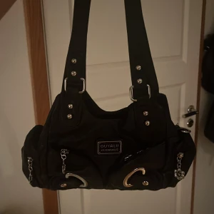 Svart axelväska från Ouyalu - Snygg svart axelväska från Ouyalu med silverdetaljer och dekorativa blommor. Väskan har flera dragkedjor och nitar som ger en cool look. Perfekt för att bära dina dagliga nödvändigheter med stil. Den är även helt oanvänd pga att jag råka beställa två.