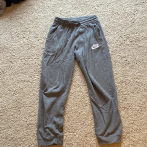 Grå mjukisbyxor från Nike - Säljer ett par grå mjukisbyxor från Nike med elastisk midja och snörning. Byxorna har sidofickor och en broderad Nike-logga på ena benet. Perfekta för en avslappnad stil. Storlek XS-S