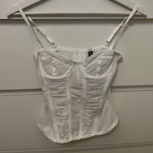 Vit korsett topp - Snygg vit korsett från H&M med justerbara axelband och dekorativa sömmar. Perfekt för att ge en stilren look. Aldrig använd ny med prislapp kvar.