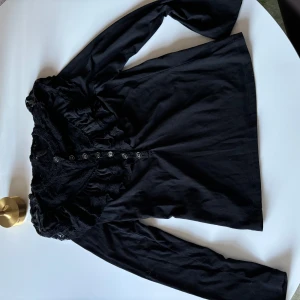 Svart spetsblus från Vero Moda - Elegant svart blus från Vero Moda med spetsdetaljer och knappar framtill. Perfekt för en stilren look med långa ärmar och vacker spetsdesign.