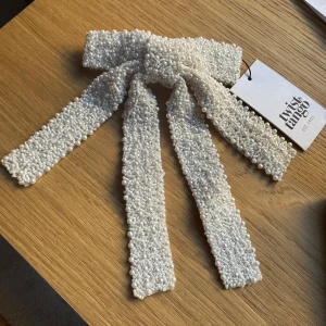 Vitt pärlhårspänne från Twist & Tango - Elegant hårspänne från Twist & Tango, dekorerat med små pärlor och en vacker rosettform. Perfekt för att ge en lyxig touch till din outfit. Helt oanvänd 
