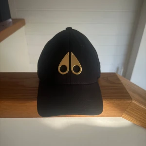 Moose Knuckles gold logo icon cap - Snygg svart moose knuckles keps med en unik gulddetalj framtill. Kepsen har en justerbar rem baktill med ett guldfärgat spänne för perfekt passform. Kepsen är köpt på NK för ca 1,5 år sedan men knappt använd. Den är i nyskick och har inga repor✨Kvitto finns!