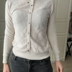 Vit ribbad kofta - Säljer en stilren vit kofta med ribbad struktur och låtsas knappar framtill (dock har de tappat lite färg men inget man märker av tydligt)