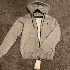 Grå hoodie från Ralph Lauren - HELT NY. Säljer en stilren grå hoodie från Ralph Lauren med dragkedja och justerbar huva. Tröjan har långa ärmar och ribbade muddar. Perfekt för en avslappnad look. STORLEK M. MEDDELA MIG INNAN KÖP