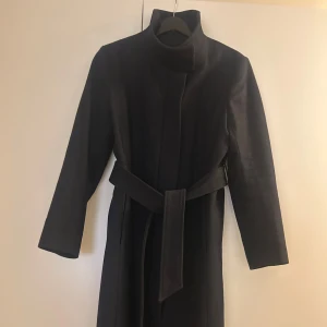 Svart kappa från Tiger of Sweden - Elegant svart kappa från Tiger of Sweden i ullblandning. Kappan har en stilren design med knytbälte i midjan och långa ärmar. 