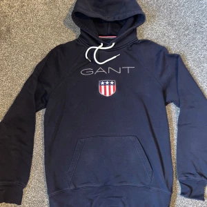 Mörkblå hoodie från GANT - Säljer en snygg mörkblå hoodie från GANT med broderad logga och emblem på bröstet. Tröjan har en justerbar huva med vita snören och en stor ficka framtill. Perfekt för en avslappnad stil.