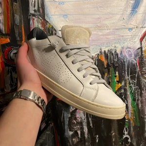 Vita sneakers från P448 - Snygga vita sneakers från P448 med perforerade detaljer och grå snörning. Skorna har en svart hälflik och är tillverkade i Italien. Perfekta för en stilren look.