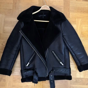 Svart skinnjacka med bälte - Snygg svart skinnjacka med dragkedja och bälte i midjan från Zara. Jackan har en klassisk design med långa ärmar och en mjuk krage. Jackan har en liten defekt där man hänger den men annars väldigt fint skick!