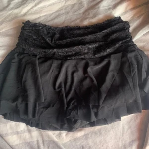 Svart kortkjol med spetsdetaljer - Snygg svart kortkjol med volang och spetsdetaljer i midjan. Perfekt för en stilren look. Kjolen har en flödande passform och är lätt att matcha med olika toppar. Köpt för 200kr endast använd en gång 