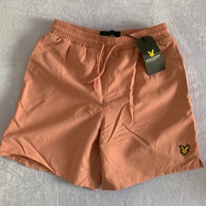Rosa badshorts från Lyle & Scott - Snygga rosa badshorts från Lyle & Scott med elastisk midja och snörning. Perfekta för sommardagar, med en praktisk bakficka och det ikoniska logotypmärket på framsidan. Helt oanvända pga fel storlek.