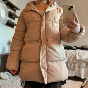 Beige dunjacka - Säljer en mysig beige pufferjacka med huva. Jackan har långa ärmar och stängs med dragkedja. Perfekt för kyliga dagar!