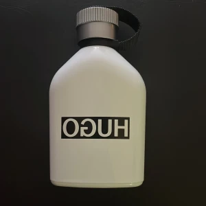 Vit parfymflaska från Hugo - Säljer en stilren vit parfymflaska från Hugo med en svart logotyp. Flaskan har en silverfärgad skruvkork och ett praktiskt svart band för enkel hantering. Perfekt för den som gillar en minimalistisk design.