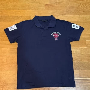 Marinblå pikétröja från Cross Sportswear - Snygg marinblå pikétröja från Cross Sportswear med tryck och broderad detalj på bröstet och ärmen. Tröjan har en klassisk krage och korta ärmar, perfekt för en sportig look.