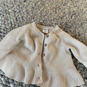 Beige stickad kofta från Polarn O. Pyret - Söt beige stickad kofta för baby från Polarn O. Pyret. Koftan har knappar framtill och en fin struktur med volangdetalj vid nederkanten. Perfekt för att hålla de små varma och stiliga.