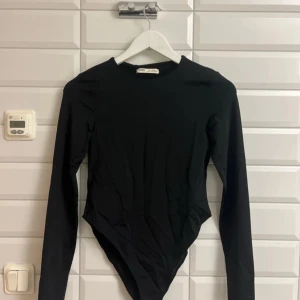 Svart långärmad topp från Pull & Bear storlek L - Snygg svart långärmad topp från Pull & Bear. Perfekt för en stilren look. Tillverkad i ett mjukt material som ger en bekväm passform. En svart helt oanvänd body ifrån pull&bear. Har aldrig använt, och inte  testat body på.  Säljer för 195 kronor. 