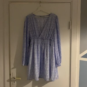 Ljusblå blommig klänning - Charmig ljusblå klänning med blommigt mönster och v-ringad design. Klänningen har långa ärmar och en smickrande omlott-look. Perfekt för vår och sommar!