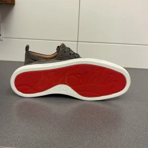 Grå sneakers från Louboutin - Snygga grå sneakers i mocka från Louboutin med den ikoniska röda sulan. Skorna har snörning och en stilren design som passar perfekt för en trendig look.