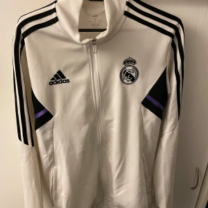 Vit träningsjacka från Adidas - Säljer en vit träningsjacka från Adidas med Real Madrid-logga. Jackan har svarta och lila detaljer samt dragkedja framtill. Perfekt för fotbollsfans!