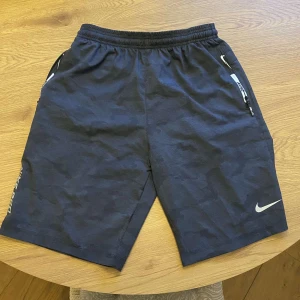Svarta shorts från Nike - Snygga svarta shorts från Nike med en diskret camouflagemönster. De har elastisk midja med snörning och dragkedja på fickorna. Perfekta för träning eller en avslappnad dag.  Nyskick, helt oanvända!