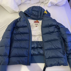 Blå dunjacka från Parajumpers - Snygg blå dunjacka från Parajumpers med huva och dragkedja. Jackan har en quiltad design och är perfekt för kyligare väder. Den har en stilren look med en liten gul detalj vid kragen.