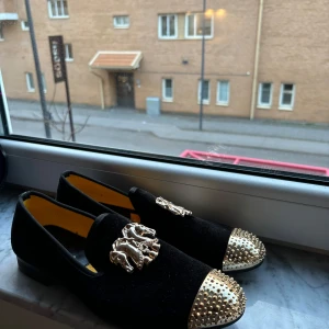 Svarta loafers med gulddetaljer från Lemax - Eleganta svarta loafers i sammet med guldfärgade metalldetaljer och nitar på tån. Skorna har en dekorativ hästdesign på ovansidan och en låg klack.