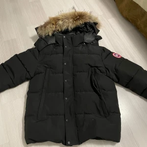 Canada goose jacka - Säljer en svart dunjacka från Canada Goose med pälskantad huva och ikonisk logga på ärmen. Jackan har en stilren design med knappar och dragkedja framtill. Perfekt för kalla vinterdagar.DET GÅR ATT TA AV PÄLSEN PÅ LUVAN