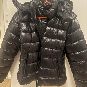 Svart pufferjacka från Elvine - Populära pufferjackan från lager 157 med dragkedja och huva. Perfekt för kyliga dagar med sin vadderade design. Jackan har praktiska fickor med dragkedjor och en glansig finish. 