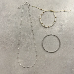 Smyckeset med halsband och armband - Elegant smyckeset med ett långt silverfärgat halsband och två armband. Ett armband är justerbart med vita och guldiga pärlor, medan det andra är ett enkelt silverfärgat armband med glitter stenar på