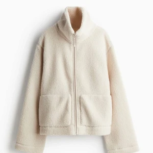 Ny naturvit / ljusbeige teddyjacka med dragkedja i M från H&M - Ny och oanvänd med tags kvar. Mysig pilejacka med hög krage och dragkedja framtill. Jackan har två stora fickor och är perfekt för kyliga dagar. Den har en avslappnad passform och långa ärmar.