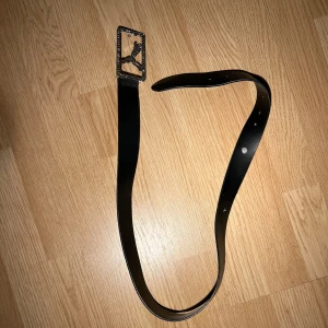 Svart bälte med rinestones spänne - Snyggt svart bälte från Puma med ett rektangulärt spänne prytt av en glittrig Puma-logga. Perfekt för att ge en sportig touch till din outfit. Bältet är gjort av skinn och har flera justerbara hål för bästa passform. Även ett eget gjort hål