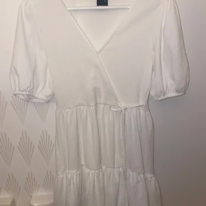 Vit omlottklänning från Gina Tricot - Säljer en söt vit omlottklänning från Gina Tricot i storlek XS. Klänningen har korta puffärmar och ett knytband i midjan för en fin passform. Perfekt för sommarens alla tillfällen!