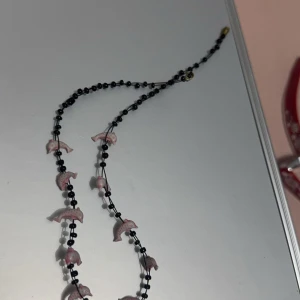 Svart halsband med rosa delfiner barn - Unikt svart halsband med små rosa delfiner som hänger längs kedjan. Perfekt för att ge en subtil färgklick till din outfit. Passar både till vardag och speciella tillfällen.