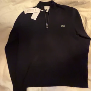 Mörkblå tröja med dragkedja från Lacoste - Säljer en stilren mörkblå tröja från Lacoste med dragkedja och det klassiska krokodilmärket på bröstet. Tröjan har en regular fit och är perfekt för en avslappnad men snygg look. Passar bra till både jeans och chinos.